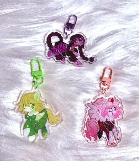 Three acrylic keychains featuring cartoon pony characters, arranged on a white faux fur background. The keychains include a green-clad character with a green clasp, a pink-haired character with a pink clasp, and a dark purple-haired character with a purple clasp.
