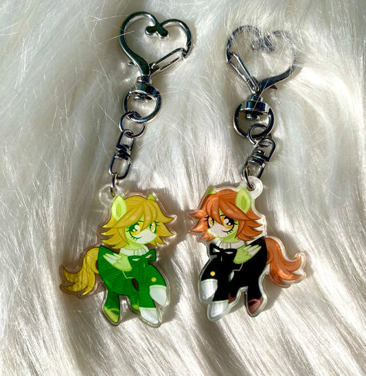 Two acrylic keychains shaped like cartoon pony characters, displayed on a white faux fur background. One keychain features a yellow-haired character in a green outfit, and the other features an orange-haired character in a black outfit.