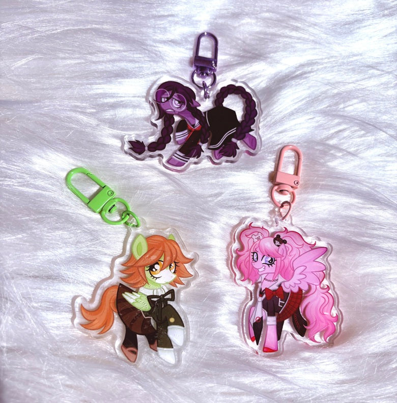 Three acrylic keychains featuring anime-style pony characters, displayed on a white faux fur background. The keychains include a purple-haired character with a silver clasp, an orange-haired character with a green clasp, and a pink-haired character with a pink clasp.