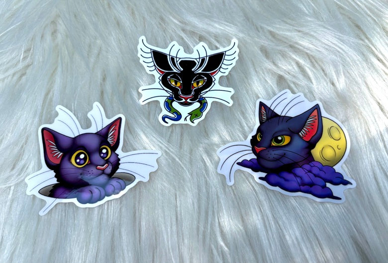 Three die-cut vinyl stickers featuring cartoon cat illustrations on a white faux fur background. The left sticker shows a purple cat peeking out from white feathers.